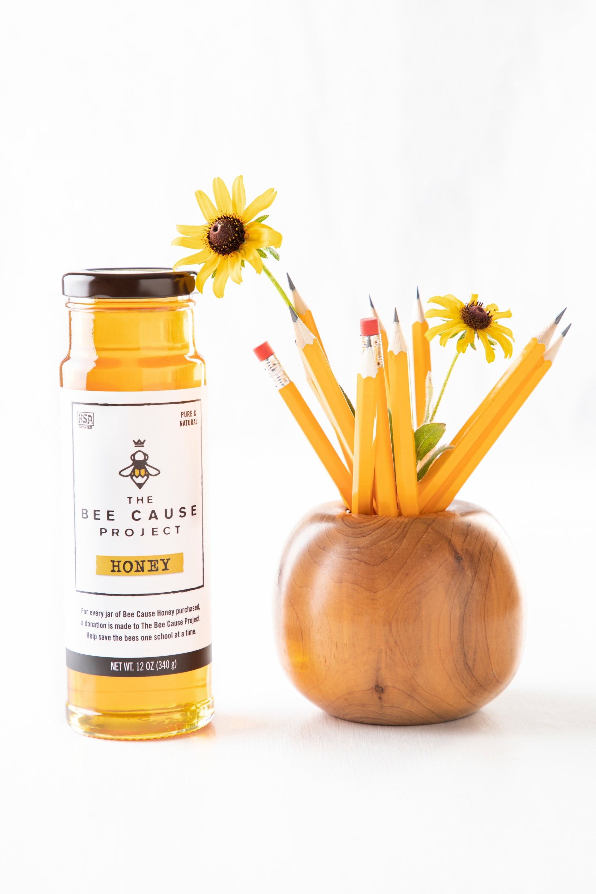 Bee Cause Honey in a 12oz tower sitting beside a wooden pencil holder, holding number 2 pencils with yellow daisy's. 
