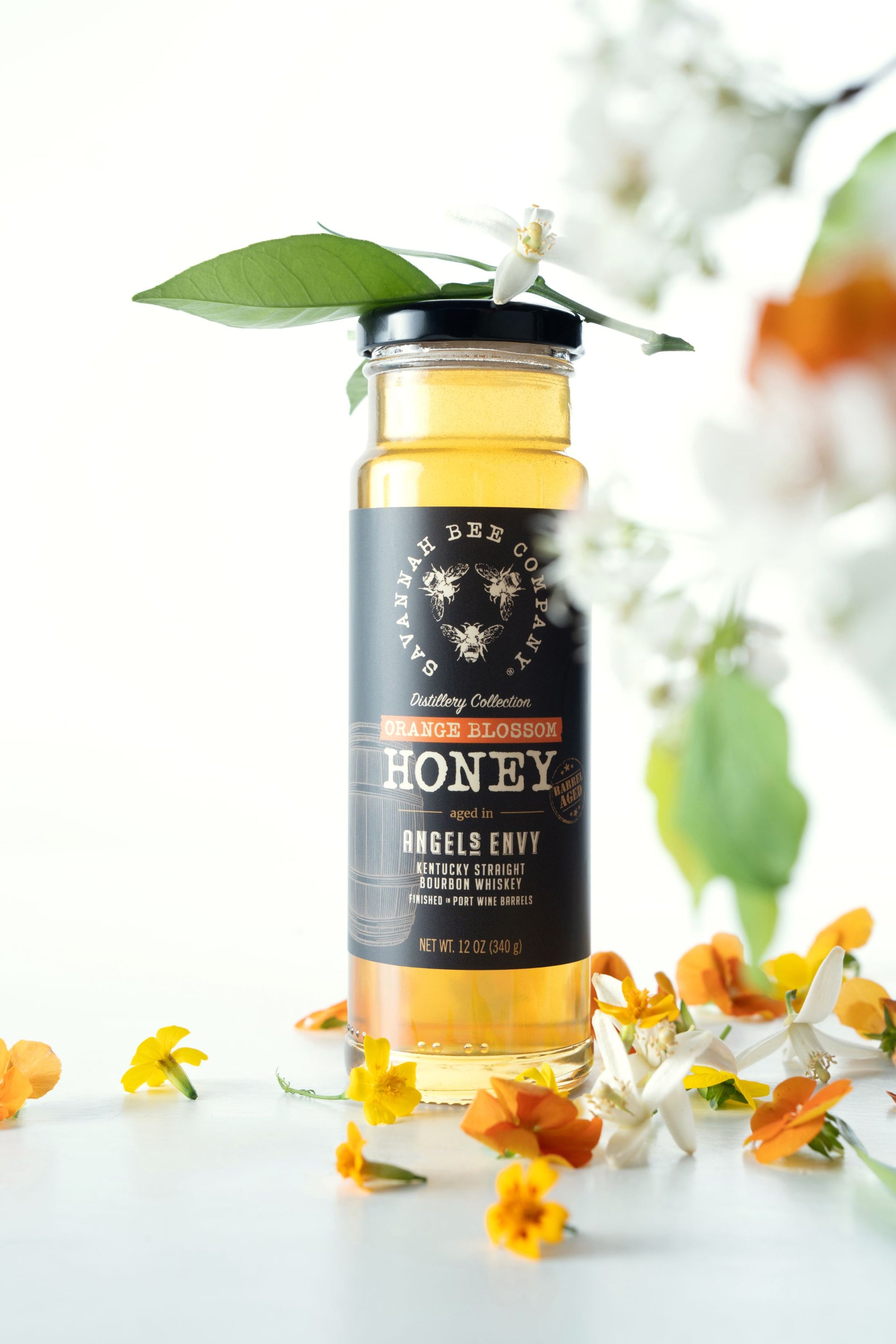 12 ounce Port Bourbon Barrel Aged Orange Blossom Honey surrounded by orange blossoms against a white background.